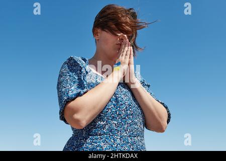 Ukrainische Frau mit einem Flaggensymbol auf der Hand betet gegen den blauen Himmel. Krieg beenden Stockfoto