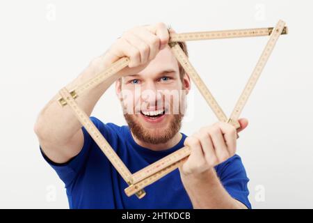 Die Maße eines Hauses. Porträt eines glücklichen jungen Mannes, der einen Tischlerherrscher in Form eines Hauses hochhält. Stockfoto