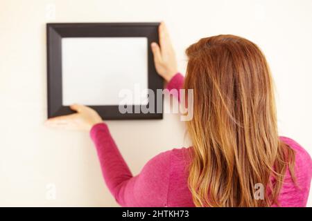 Perfekt im Bild. Rückansicht einer Frau, die einen leeren Rahmen an eine Wand legt. Stockfoto