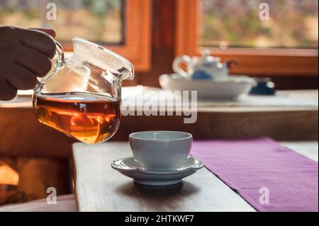 Man gießt heißen Tee aus einer Glaskanne in eine Teetasse auf einen Holztisch Stockfoto