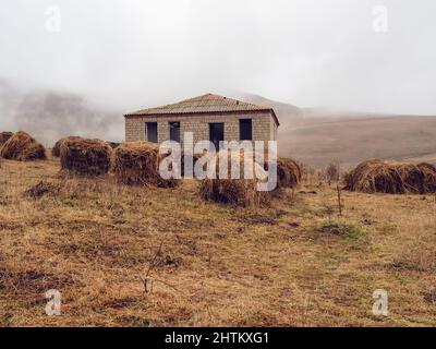 Haystacks umgeben verlassene einstöckige Backsteinhäuser auf einem Feld im Hochland, das von Nebel bedeckt ist. Stockfoto