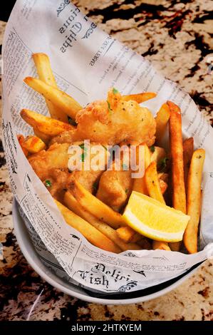 Fisch und Chips mit Zitronenkeil, eingewickelt in bedrucktes Papier auf einer Marmorstange. Stockfoto