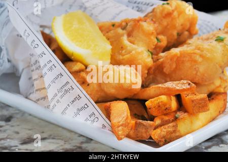 Fisch und Chips mit Zitronenkeil, eingewickelt in bedrucktes Papier auf einer Marmorstange. Stockfoto