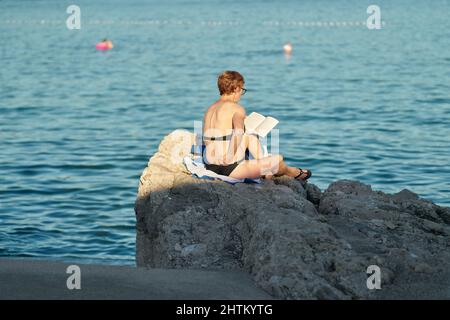 Eine Frau sitzt in der Nähe von Krk an der Küste der kroatischen Adria und liest ein Buch Stockfoto