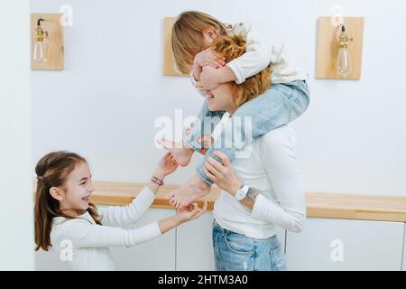 Lachende Schwester kitzelt den kleinen Jungen auf den Füßen, er sitzt auf den Schultern seiner Mutter. Im Flur. Er bedeckt die Augen seiner Mutter mit seinen Händen. Stockfoto