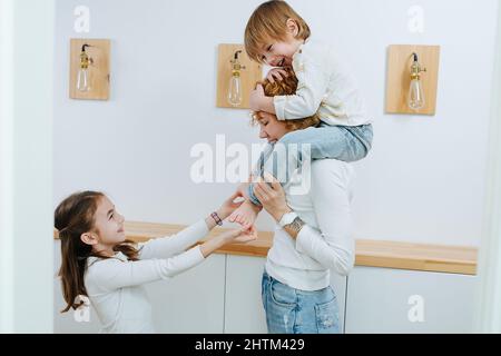 Schwester kitzelt dem kleinen Jungen die Füße, während er auf den Schultern seiner Mutter sitzt. Im Flur. Stockfoto