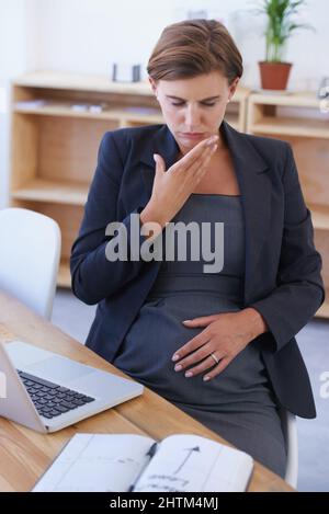Kämpfen durch die Übelkeit. Eine schwanger Geschäftsfrau, die an ihrem Schreibtisch mit Übelkeit kämpft. Stockfoto