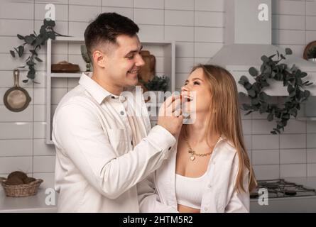 Glücklicher Mann, der Frau beim gemeinsamen Kochen in der modernen weißen scandi-Küche füttert. Romantisches Abendessen, gemeinsame Zeit. Hochwertige Fotos Stockfoto