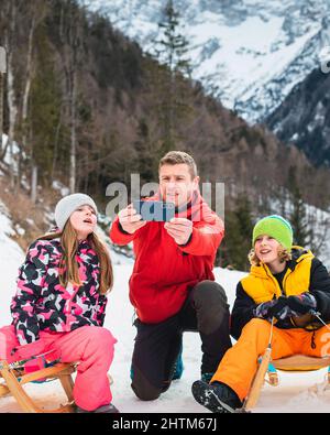 Vater mit Kindern macht Selfies, während er auf Holzschlitten sitzt, mit einer wunderschönen Berglandschaft im Hintergrund. Stockfoto