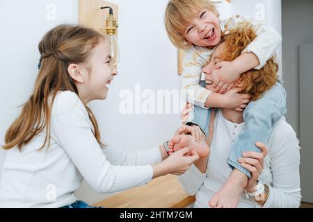 Glückliche Schwester kitzelt den kleinen Jungen auf den Füßen, er sitzt auf den Schultern seiner Mutter. Im Flur. Er hält den Kopf seiner Mutter mit den Händen. Stockfoto