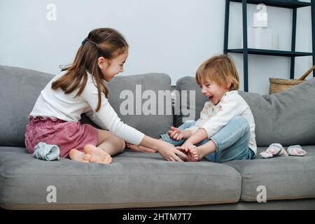 Glückliches Mädchen, das versucht, den Fuß des Jungen zu kitzeln, lachen beide von Herzen. Auf einer grauen Couch im Wohnzimmer. Stockfoto