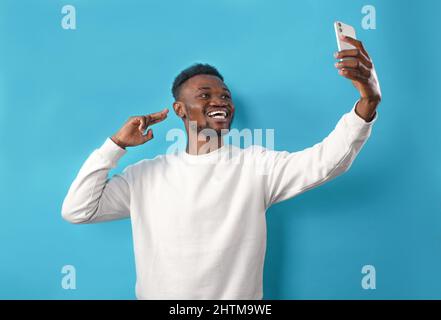 Porträt eines afroamerikanischen Mannes, der ein Selfie macht und auf einem blau isolierten Hintergrund lächelt Stockfoto