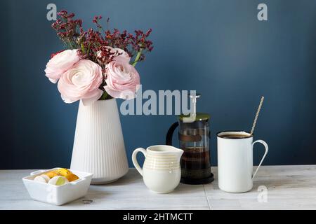 Kaffeetasse und Bouquet von rosa persischen Butterblumen auf Holztisch. Mock up Hintergrund für Design. Gelbe und weiße Makronen in einer weißen Porzellanschale Stockfoto