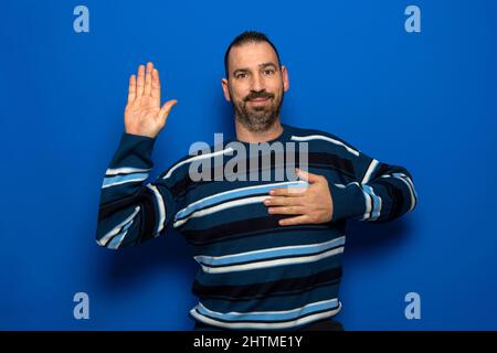 Ein junger, hübscher Mann mit Bart trägt einen lässigen Pullover auf blauem Hintergrund und schwört mit der Hand auf der Brust und der offenen Handfläche, was einen Treueversprechen-Eid ablegt Stockfoto