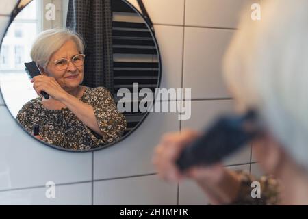 Ältere kaukasische Frau kämmt sich die Haare und schaut auf den Spiegel. Hochwertige Fotos Stockfoto