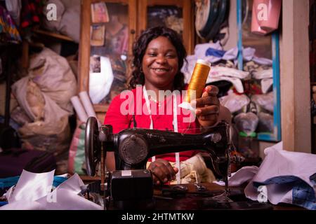 Eine afrikanische Näherin hält einen Bobbin in der Hand und arbeitet in ihrem Schneidergeschäft, sie ist glücklich und lächelt Stockfoto
