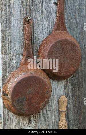 Alte verrostete Bratpfannen hängen an der grau verwitterten Außenwand einer alten rustikalen Holzplankenhütte. Stockfoto