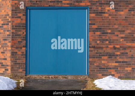 Zwei massive, blaue Metalltüren im Eingangsbereich eines rot-braunen Gebäudes. Die Türen haben keine Griffe und werden nur von innen geöffnet. Stockfoto