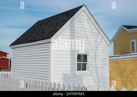 Ein kleiner weißer Holzschuppen mit einem einzigen Fenster in der Mitte und schwarzen Schindeln auf dem Dach in einem Garten mit einem weißen Zaun. Stockfoto