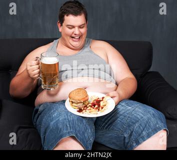 Etwas, um sein Essen abzuwaschen. Ein übergewichtiger junger Mann, der auf einem Sofa sitzt und sich an Bier und Fast Food trinkt. Stockfoto