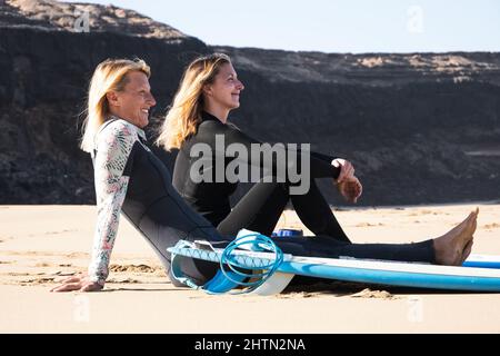 2 kaukasische Surfer sitzen am Strand. Sie lächeln zum Meer. Sport, gesunder Lebensstil, Freundschaftskonzept. Stockfoto