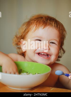 Lustige Baby Essen selbst mit einem Löffel in der Küche. Lächelndes Baby beim Essen. Stockfoto