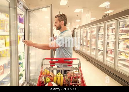 Glücklicher Mann mit Einkaufswagen. Kunde Verbraucher mit Einkäufen. Kerl im Lebensmittelgeschäft. Stockfoto