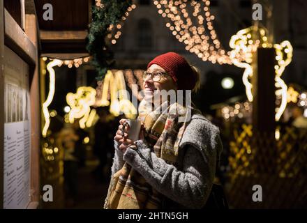 Junge Frau hält den Becher am geschmückten Stand auf dem Weihnachtsmarkt Stockfoto