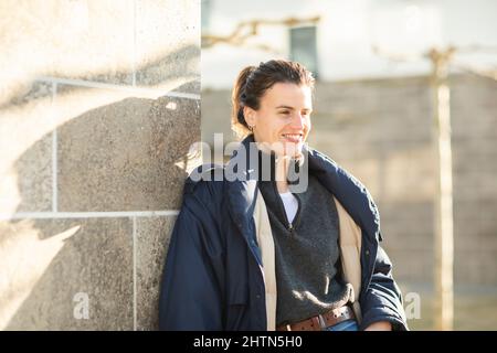 Lächelnde Frau in warmer Jacke, die sich an die Wand lehnt Stockfoto