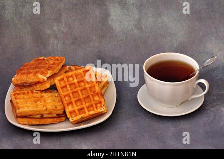 Eine Tasse schwarzen Tee und ein Teller mit mehreren frischen Wiener Waffeln auf einem grauen abstrakten Hintergrund. Nahaufnahme. Stockfoto