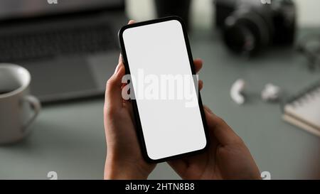 Ausgeschnittene Aufnahme einer jungen Beamtin, die ein Mobiltelefon mit leerem Bildschirm über ihren Arbeitstisch hält. Stockfoto