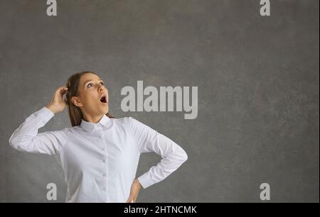Fröhliche Frau mit einem überraschenden Ausdruck schaut auf, während sie auf einem grauen Hintergrund steht. Stockfoto