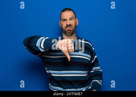 Junger, hübscher Mann mit gestreiftem Pullover, der auf einem isolierten blauen Hintergrund steht und unglücklich und wütend aussagt und Ablehnung und negative Geste zeigt Stockfoto