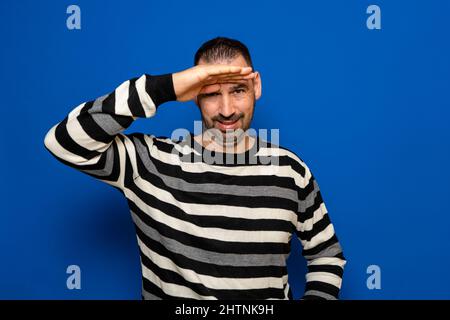 Der Mann sucht etwas. Der wachsame Mann kämpft darum, aus der Ferne zu sehen. Er trägt einen gestreiften Pullover. Isoliert auf blauem Hintergrund. Stockfoto