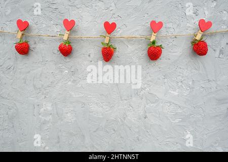 Girlande aus roten reifen Erdbeeren, die auf Holzkeilspinnchen mit Herzen auf Seilgarn hängen, am oberen Rand auf grauem Hintergrund. Kopierbereich Creative t Stockfoto