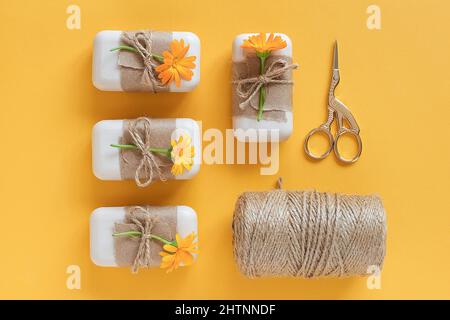 Handgemachte Naturseife, verziert mit Bastelpapier, orangen Ringelblumen, Garnstrang und Schere auf gelbem Hintergrund. Bio-Kosmetik Co Stockfoto