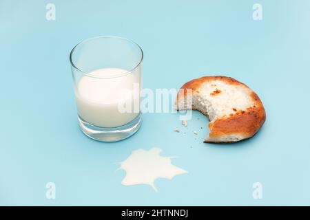 Halb Glas Milch, frisch gebackenes Brötchen mit Quark, verschüttete Milch und Krümel auf blauem Hintergrund abgebissen. Konzept nahrhaftes Frühstück oder Snacks Stockfoto