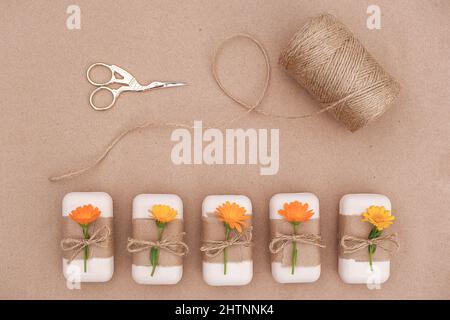 Handgemachte Naturseife mit Bastelpapier, orangen Ringelblumen, Garnklee und Schere verziert. Bio-Kosmetik-Konzept. Draufsicht flach Stockfoto