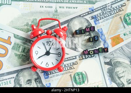 Zeit ist Geld Text und roter Wecker auf dem Hintergrund der amerikanischen Hunderte Dollar-Scheine, Nahaufnahme. Kreatives Konzept Zitat des Tages. Draufsicht Flat Lay Stockfoto
