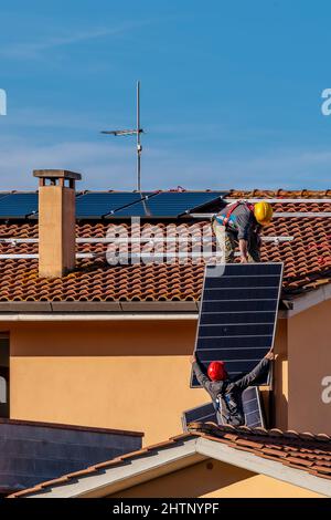 Zwei Arbeiter führen Solarzellen aneinander vorbei, um auf einem roten Ziegeldach montiert zu werden, um eine Photovoltaikanlage zu installieren Stockfoto