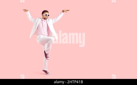 Lustiger Mann in weißem Anzug und Sonnenbrille, der auf einem Fuß auf einem rosa Kopierraum-Hintergrund steht Stockfoto