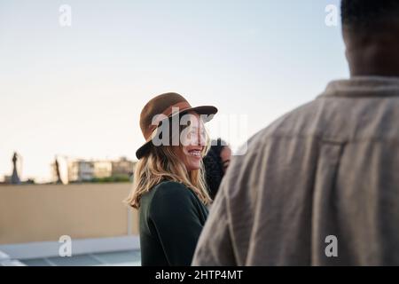 Kaukasische Frau lächelt und spricht mit einer Gruppe von Freunden, die auf dem Dach bei einer Party in der Stadt zusammenstehen Stockfoto