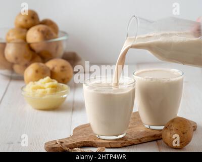 Kartoffelmilch, die auf weißem Holzhintergrund in Glas gießt. Vegane Milch in Glas gießen, mit Kartoffelpüree und Kartoffelknollen auf dem Hintergrund. Speicherplatz kopieren. Hausgemachte Kartoffelmilch aus gekochten Kartoffeln Stockfoto