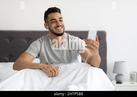 Online-Kommunikation. Glücklich Arabischen Mann Mit Smartphone Im Bett Für Videoanruf Stockfoto