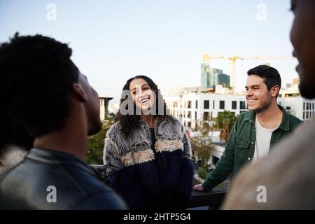 Biracial Frau lächelt mit einer Gruppe von multikulturellen Freunden, die sich in der Abenddämmerung auf einer Dachterrasse treffen. Stockfoto