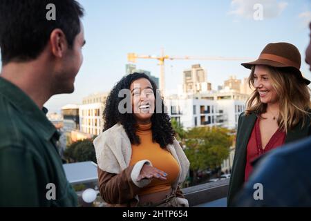 Biracial Frau lachend mit Gruppe von multikulturellen Freunden gesellig auf einer Dachterrasse in der Dämmerung. Stockfoto