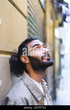 Bärtiger hispanischer Mann in legerer Kleidung mit Pferdeschwanz, der nach oben schaut und träumt, während er sich an einer Wand auf der Stadtstraße lehnt Stockfoto
