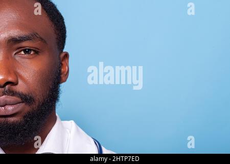 Zugeschnittenes Bild eines afroamerikanischen Mediziners, der selbstbewusst und entspannt in weißem Labormantel mit Stethoskop auf blauem Hintergrund aussieht. Nahaufnahme des halben Gesichts Porträt eines Arztes mit braunen Augen. Stockfoto