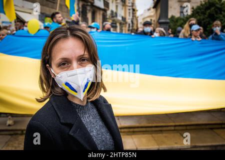 Trauriges ukrainisches Mädchen mit Gesichtsmaske, gemalt in den Farben der Ukraine und einer großen Nationalflagge dahinter Stockfoto