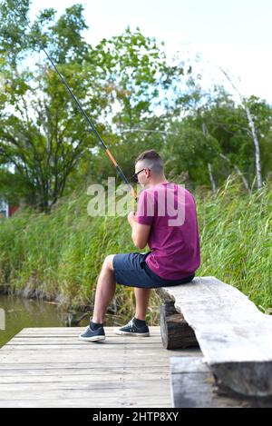 Junger Fischer sitzt auf hölzernen Pier, Angeln im See. Er hält die Rute und schaut auf Schwimmer Stockfoto